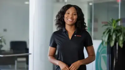 A smiling young Black woman in a black polo shirt with a logo stands in a bright, modern office space.